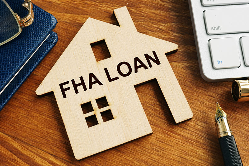 A wooden house-shaped cutout with FHA LOAN written on it sits on a wooden desk next to a notebook, glasses, a keyboard, and a fountain pen.